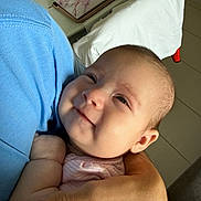 Lucia is registered to the contest to win money with this photo: baby, infant, smile, person, arm, hand, holding, cradle, cheek, portrait, indoor, blue_shirt, pink_clothing, pillow, table, charger, floral, tile_floor, ear, closeup