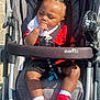 Omri is registered to the contest to win money with this photo: baby, stroller, infant, child, red_shoes, hand_on_mouth, casual_clothing, sunlight, outdoor, seatbelt, pacifier, black_hair, curly_hair, fabric, footwear, baby_bib, resting, cute, small_person, daylight