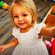 Gracelynn is registered to the contest to win money with this photo: child, toddler, smiling, white_dress, blonde_hair, indoor, wooden_floor, toy, furniture, person, happy, portrait, cute, young, girl, home, flooring, playroom, childhood, innocence
