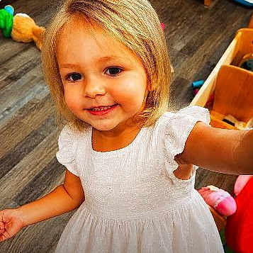 Gracelynn is registered to the contest to win money with this photo: child, toddler, smiling, white_dress, blonde_hair, indoor, wooden_floor, toy, furniture, person, happy, portrait, cute, young, girl, home, flooring, playroom, childhood, innocence