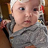baby, infant, child, portrait, close_up, wide_eyes, headband, gray_outfit, clothing, sitting, held, adult_hand, indoor, home, wood_cabinet, furniture, warm_lighting, candid, face, photography