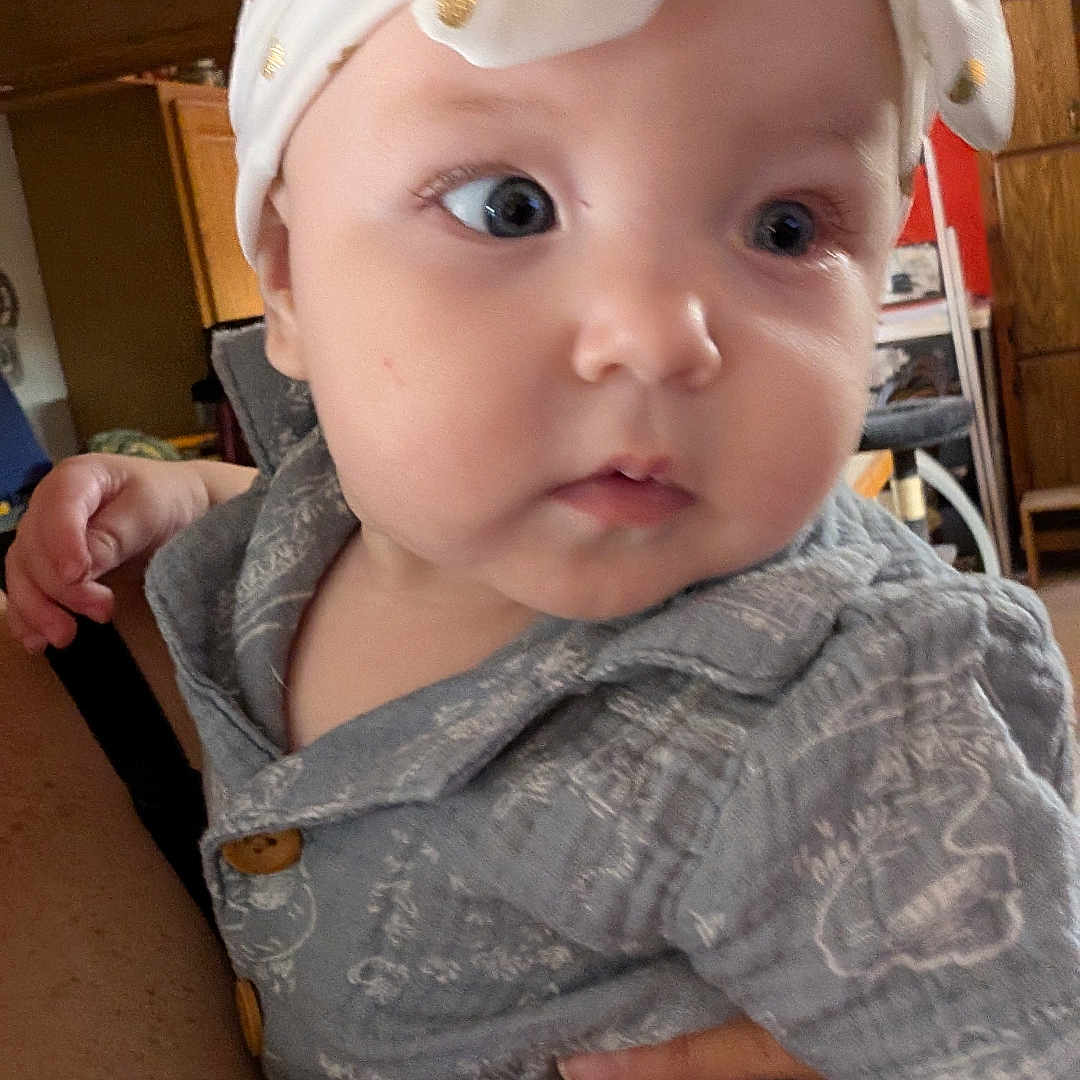 Blair joined the competition — help win amazing prizes! baby, infant, child, portrait, close_up, wide_eyes, headband, gray_outfit, clothing, sitting, held, adult_hand, indoor, home, wood_cabinet, furniture, warm_lighting, candid, face, photography