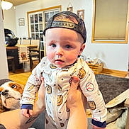 Kole is registered to the contest to win money with this photo: baby, child, person, dog, cap, baseball, pajamas, tattoo, hand, living_room, carpet, furniture, window, curtains, table, chair, lamp, dog_face, cute, indoor