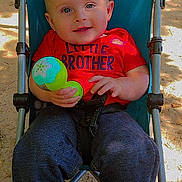 Noah is registered to the contest to win money with this photo: baby, child, stroller, toy, red_shirt, blue_stroller, outdoor, sand, sunlight, smiling, happy, playing, casual_clothing, infant, person, cute, portrait, daylight, relaxed, young