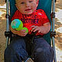 baby, child, stroller, toy, red_shirt, blue_stroller, outdoor, sand, sunlight, smiling, happy, playing, casual_clothing, infant, person, cute, portrait, daylight, relaxed, young