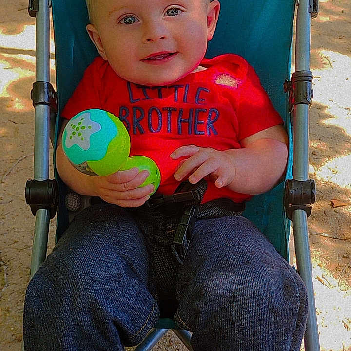Noah is registered to the contest to win money with this photo: baby, blue_stroller, casual_clothing, child, cute, daylight, happy, infant, outdoor, person, playing, portrait, red_shirt, relaxed, sand, smiling, stroller, sunlight, toy, young
