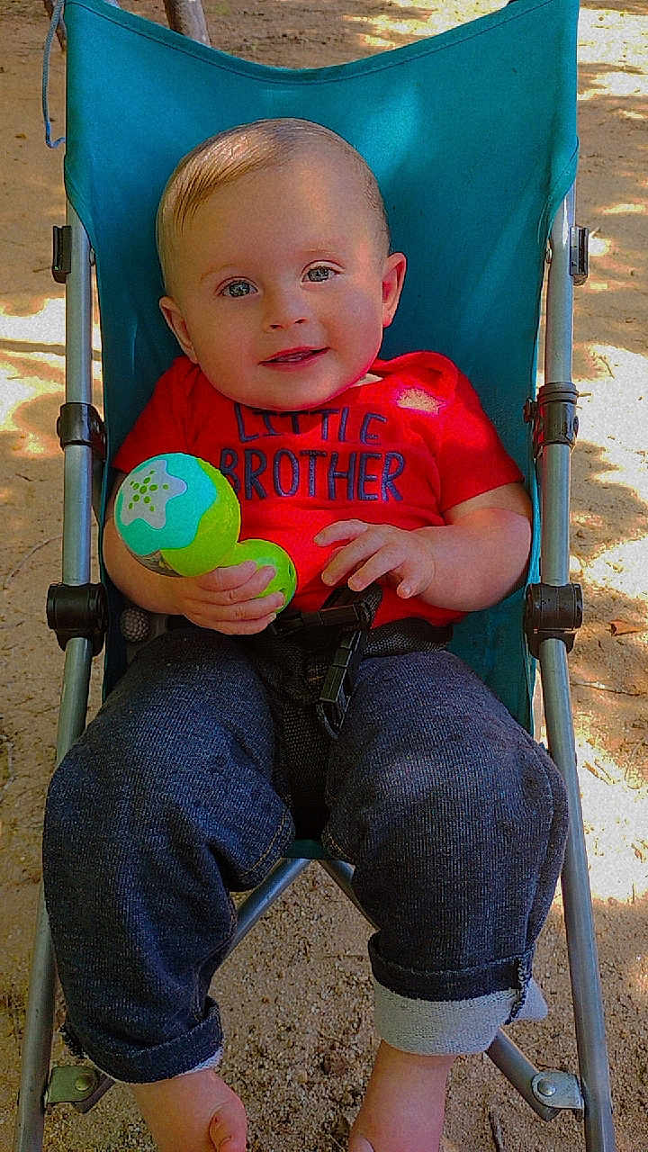 Noah is registered to the contest to win money with this photo: baby, child, stroller, toy, red_shirt, blue_stroller, outdoor, sand, sunlight, smiling, happy, playing, casual_clothing, infant, person, cute, portrait, daylight, relaxed, young