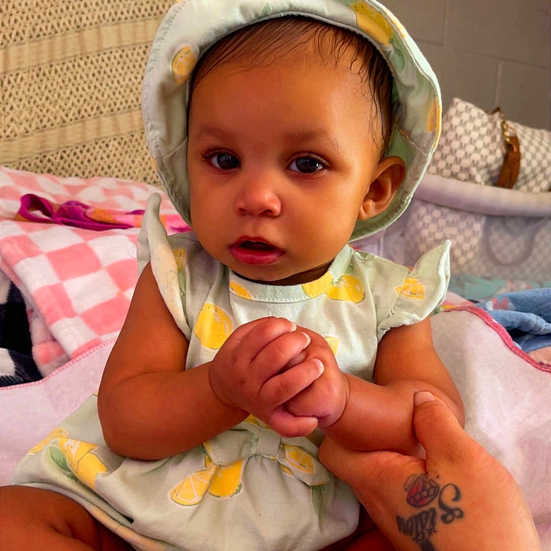 Saniah joined the competition — help win amazing prizes! baby, infant, child, holding_hand, hat, clothing, blanket, tattoo, hand, feet, indoors, seated, cute, portrait, person, soft_lighting, fabric, pattern, home, support