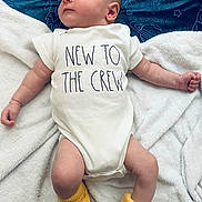 Ames is registered to the contest to win money with this photo: baby, infant, onesie, yellow_booties, blanket, space_pattern, soft_texture, cute, newborn, clothing, knitted_booties, face, lying_down, indoors, child, toddler, expression, cozy, white, casual