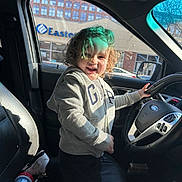 Johmyer is registered to the contest to win money with this photo: child, curly_hair, gray_hoodie, car_interior, steering_wheel, kneeling, smiling, sunlight, window, seat, sneakers, sippy_cup, toy, playful, happy, person, vehicle, casual_clothing, daylight, front_seat