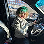 car_interior, casual_clothing, child, curly_hair, daylight, front_seat, gray_hoodie, happy, kneeling, person, playful, seat, sippy_cup, smiling, sneakers, steering_wheel, sunlight, toy, vehicle, window