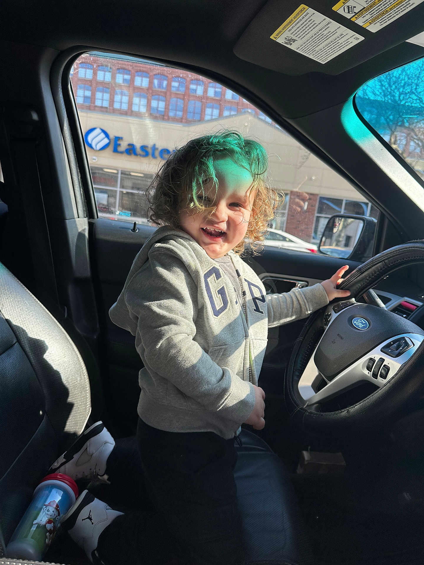 Johmyer is registered to the contest to win money with this photo: child, curly_hair, gray_hoodie, car_interior, steering_wheel, kneeling, smiling, sunlight, window, seat, sneakers, sippy_cup, toy, playful, happy, person, vehicle, casual_clothing, daylight, front_seat