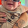 Jesse is registered to the contest to win money with this photo: baby, child, sunglasses, smiling, tattoo, arm, person, indoor, clothing, face, happy, holding, casual, portrait, cute, infant, pattern, window, sunlight, hand