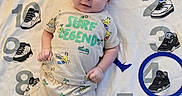 Jesse is registered to the contest to win money with this photo: baby, infant, child, cute, smiling, clothing, tshirt, shorts, lying_down, blanket, numbers, sneakers, cartoon, pattern, playful, indoors, portrait, person, happy, bright
