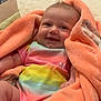 baby, infant, blanket, orange_blanket, colorful_clothing, tie_dye, smiling, happy, cute, face, indoors, soft_texture, cushion, floor, wooden_floor, child, person, young_child, comfortable, cozy