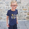child, boy, glasses, smiling, tshirt, shorts, sandals, stone_wall, pavement, window, outdoor, person, casual_clothing, summer, happy, standing, young, portrait, fashion, daylight