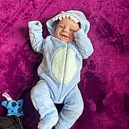 Emilio participe au concours pour gagner de l'argent avec cette photo : baby, child, onesie, blue_clothing, hood, smile, lying_down, plush_blanket, purple_background, stuffed_toy, cute, infant, soft_texture, happy, indoor, portrait, cozy, small_toy, fabric, casual