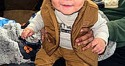 Noam is registered to the contest to win money with this photo: toddler, child, smiling, sneakers, brown_clothing, vest, adult_hand, indoor, couch, blanket, person, happy, face, hair, sitting, hoodie, casual, portrait, cozy, home