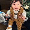 toddler, child, smiling, sneakers, brown_clothing, vest, adult_hand, indoor, couch, blanket, person, happy, face, hair, sitting, hoodie, casual, portrait, cozy, home