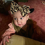 River is registered to the contest to win money with this photo: baby, child, leopard_print, bow, headwear, pink_clothing, fur_vest, hand, indoor, carpet, curious, wide_eyes, face, person, soft_light, expression, candid, portrait, infant, warm_colors