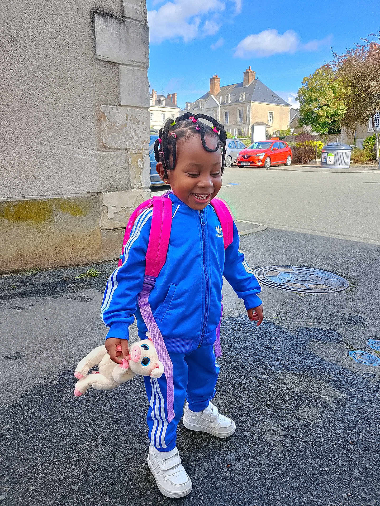 Romellia participe au concours pour gagner de l'argent avec cette photo : child, smiling, blue_tracksuit, white_sneakers, plush_toy, backpack, outdoor, street, happy, laughing, hair_ties, asphalt, building, car, daylight, urban, person, playful, cute, fashion