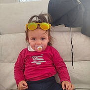 Elyna participe au concours pour gagner de l'argent avec cette photo : toddler, child, pacifier, sunglasses, pink_shirt, pants, white_couch, indoor, person, hair, cute, sitting, casual_clothing, face, young_child, living_room, window, black_jacket, expression, home