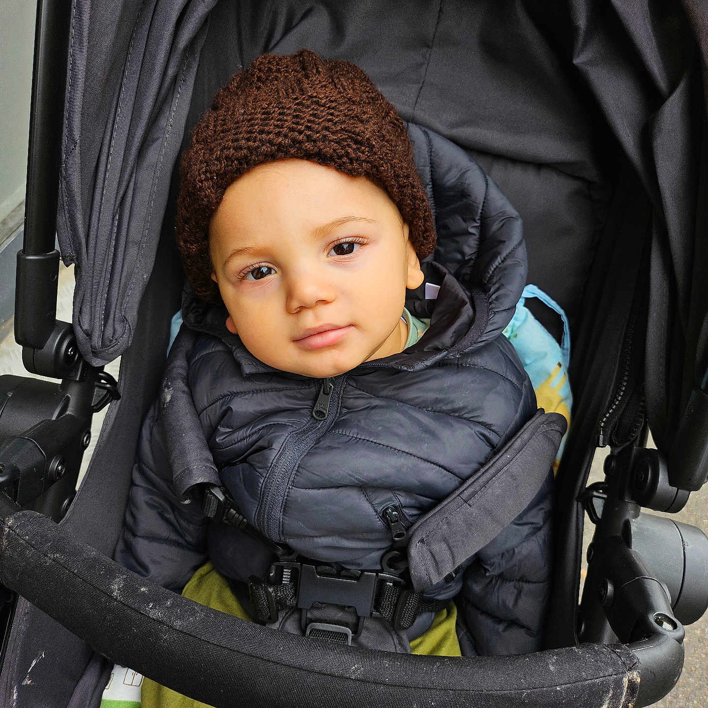 Chris-Emmanuel a rejoint le concours — aidez-le/la à gagner de superbes lots ! baby, casual, child, clothing, curious_expression, face, hat, infant, jacket, knit_hat, looking_at_camera, outdoor, person, puffy_jacket, safety_belt, seated, stroller, toddler, warm_clothes, young_child