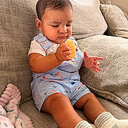 Larissa joined the competition — help win amazing prizes! baby, child, couch, overalls, socks, blanket, indoor, curious, food, snack, person, furniture, seated, portrait, skin, head, hand, expression, clothing, home