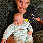 Floyd is registered to the contest to win money with this photo: baby, infant, man, father, couch, living_room, smile, happy, portrait, sitting, t_shirt, facial_hair, wood_paneling, cozy, domestic, parent_child, fabric, relaxed, cuteness, home_interior