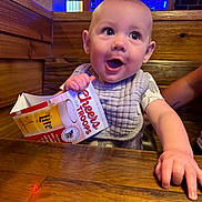 Westyn is registered to the contest to win money with this photo: baby, child, table, wood, indoor, bib, cardboard, beer_holder, text, neon_light, blue_light, hand, smiling, happy, face, curious, seated, restaurant, person, casual