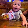 baby, child, table, wood, indoor, bib, cardboard, beer_holder, text, neon_light, blue_light, hand, smiling, happy, face, curious, seated, restaurant, person, casual