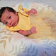 Braylee is registered to the contest to win money with this photo: baby, infant, yellow_dress, tulle_skirt, pillow, floral_pattern, newborn, cute, small_hands, lying_down, indoors, soft_lighting, portrait, child, person, sleeping_area, bedroom, resting, curious, comfortable