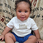 J'Khari Cruse is registered to the contest to win money with this photo: baby, child, smiling, sitting, tshirt, blue_shorts, socks, curly_hair, blanket, cozy, portrait, indoors, cute, happy, infant, person, face, young_child, warm, comfortable