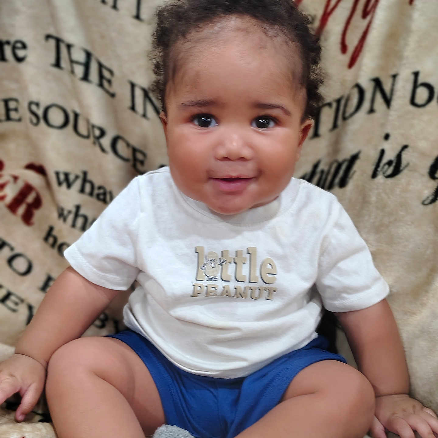 J'Khari Cruse is registered to the contest to win money with this photo: baby, child, smiling, sitting, tshirt, blue_shorts, socks, curly_hair, blanket, cozy, portrait, indoors, cute, happy, infant, person, face, young_child, warm, comfortable