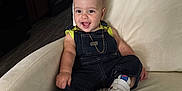 Christopher is registered to the contest to win money with this photo: baby, smiling, denim_overalls, sneakers, yellow_shirt, couch, indoor, child, happy, gold_chain, sitting, face, person, footwear, furniture, home, kitchen, floor, light, casual