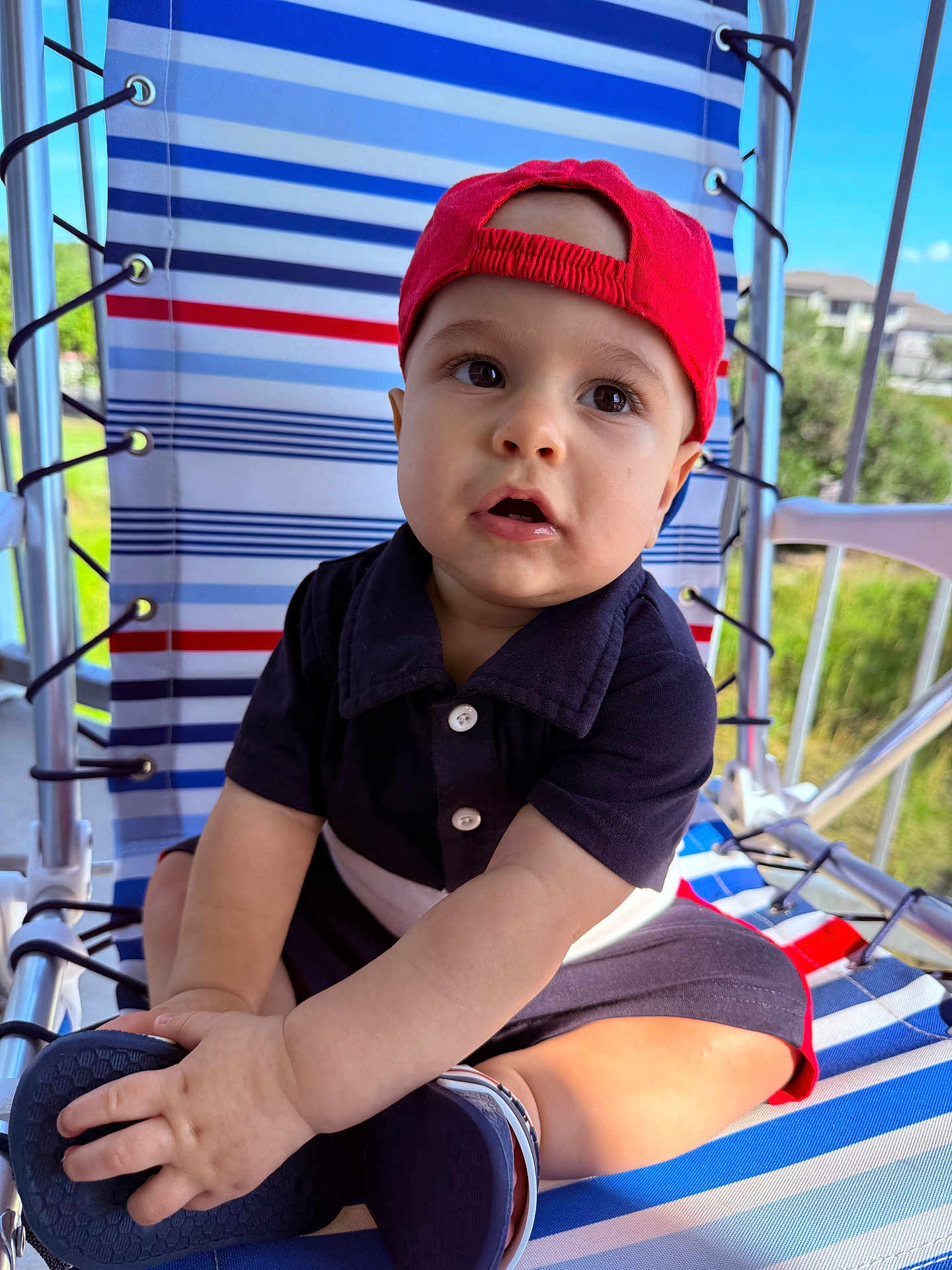 Christopher is registered to the contest to win money with this photo: toddler, child, baby, cap, red_cap, navy_outfit, shoes, striped_chair, outdoor, daylight, porch, hand, face, sitting, curious, expression, summer, greenery, background, sunlight