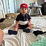 baby, child, red_cap, sitting, blanket, carpet, dog, baby_walker, indoor, floor, shoe, bottle, pillow, toys, person, home, casual_clothing, curious, young_child, relaxed