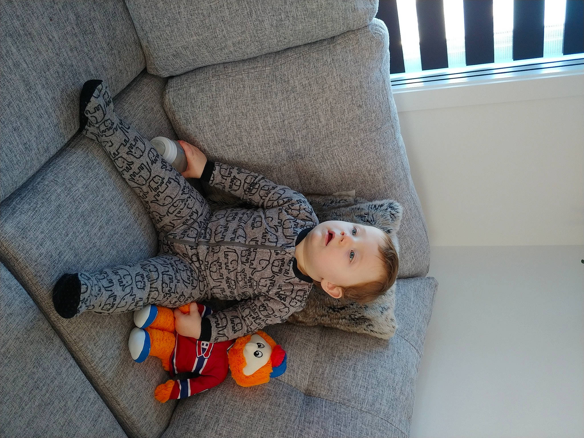 Liam a rejoint le concours — aidez-le/la à gagner de superbes lots ! baby, carmine, child, comfort, doll, floor, flooring, grey, human_leg, leg, linens, pattern, person, room, sitting, stuffed_toy, toddler, toy, window_blind, wood