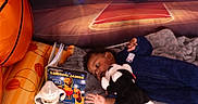 Akeem is registered to the contest to win money with this photo: baby, infant, blanket, stuffed_animal, toy, book, bottle, basketball, inflatable, sports_theme, child, floor, indoor, blue_clothing, soft_texture, lying_down, reading, black_hair, nursery, playtime