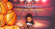 Akeem is registered to the contest to win money with this photo: background, basketball, black_hair, book, child, clothing, court, floor, indoor, inflatable, lights, playful, portrait, reading, seats, shoes, smiling, sports, stadium, toddler