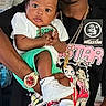 Akeem is registered to the contest to win money with this photo: baby, child, person, adult, holding, white_shoes, green_shorts, white_tshirt, black_tshirt, gold_chain, watch, cartoon_graphic, indoor, portrait, face, hand, cute, fashion, accessory, expression