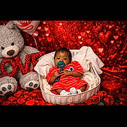 Akeem is registered to the contest to win money with this photo: baby, basket, teddy_bear, valentines_day, heart_shaped, balloon, pacifier, red_clothing, roses, decorations, cute, infant, portrait, indoor, holiday_theme, love, plush_toy, seated, background, smiling
