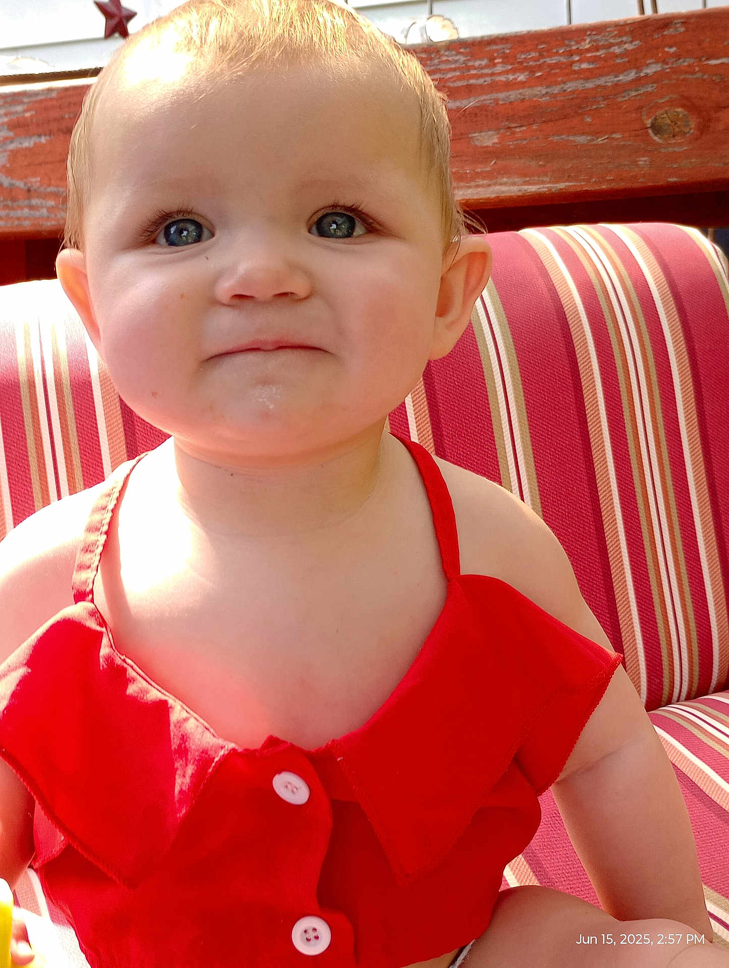 Freya is registered to the contest to win money with this photo: toddler, child, face, blue_eyes, red_clothing, smile, sitting, cushion, striped_pattern, wooden_background, sunlight, portrait, cute, young_child, baby, outdoor, happy, skin, buttoned_top, person