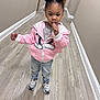 child, toddler, pink_jacket, jeans, sneakers, hair_buns, finger_in_mouth, necklace, hallway, wood_floor, baseboard, beige_wall, door, standing, portrait, indoor, cute, casual_photo, young_child, expression
