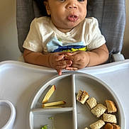 Adyn is registered to the contest to win money with this photo: child, toddler, high_chair, divided_plate, apple_slices, sandwich_cubes, broccoli, hands, messy_face, drooling, curly_hair, white_shirt, tray, feeding, mealtime, indoor, portrait, close_up, baby_food, expression