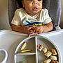 child, toddler, high_chair, divided_plate, apple_slices, sandwich_cubes, broccoli, hands, messy_face, drooling, curly_hair, white_shirt, tray, feeding, mealtime, indoor, portrait, close_up, baby_food, expression