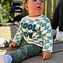 toddler, child, sitting, outdoor, wooden_deck, green_pants, white_shoes, checkered_sweater, curious, sunlight, person, blurred_background, stroller, adult, casual_clothing, daylight, portrait, expression, baby, cute