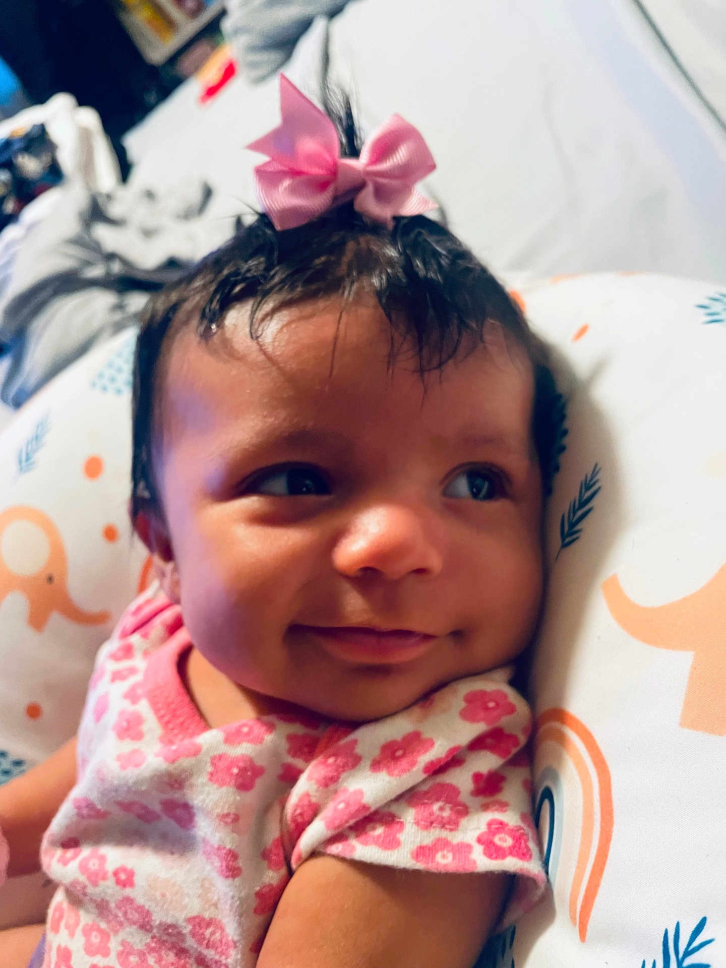 Gianna is registered to the contest to win money with this photo: baby, child, smile, pink_bow, floral_clothing, cushion, elephant_pattern, rainbow_pattern, face, portrait, indoors, cute, infant, resting, soft_lighting, closeup, cheeks, happy, young_child, headwear