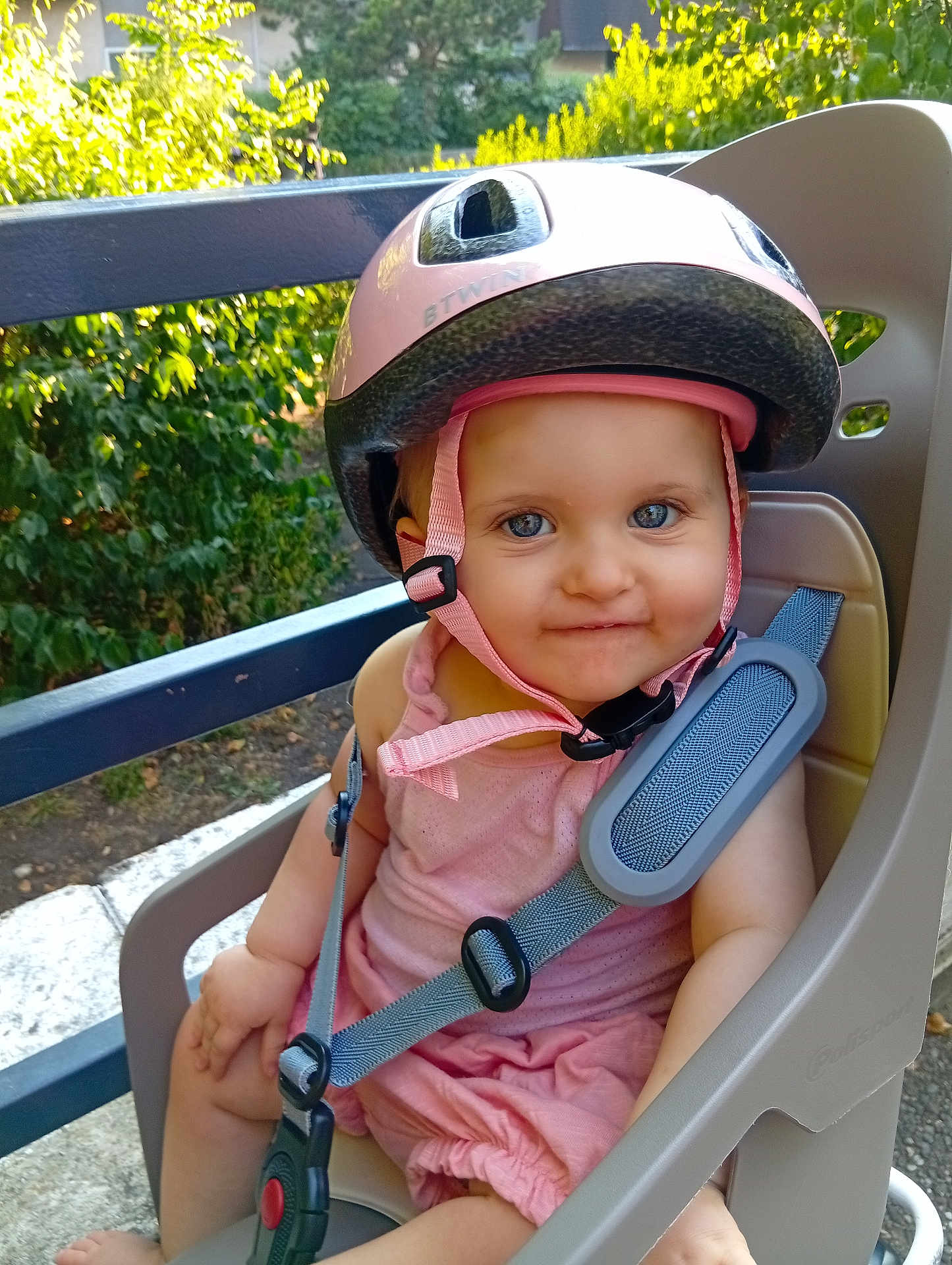 Helena participe au concours pour gagner de l'argent avec cette photo : baby, child, helmet, pink, blue_eyes, smile, child_bike_seat, straps, outdoor, greenery, safety, seat, person, summer, cute, infant, sunlight, headgear, happy, casual