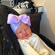 Delaine is registered to the contest to win money with this photo: baby, infant, smiling, headband, bow, blanket, clothing, face, sleeping, person, indoors, furniture, table, fabric, black, purple, cozy, cute, resting, portrait